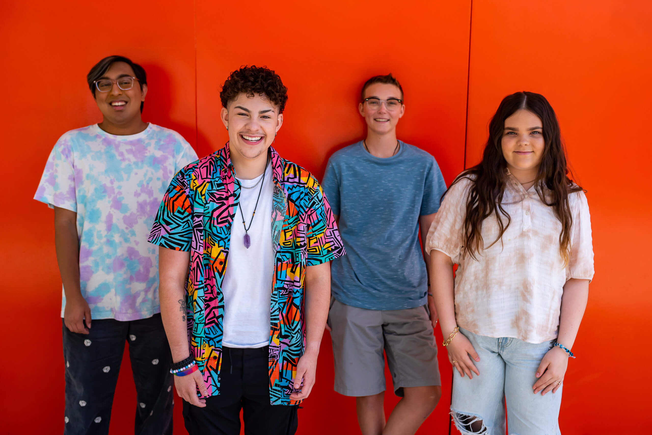 A diverse group of four LGBTQ+ young people smile in front of an orange wall.