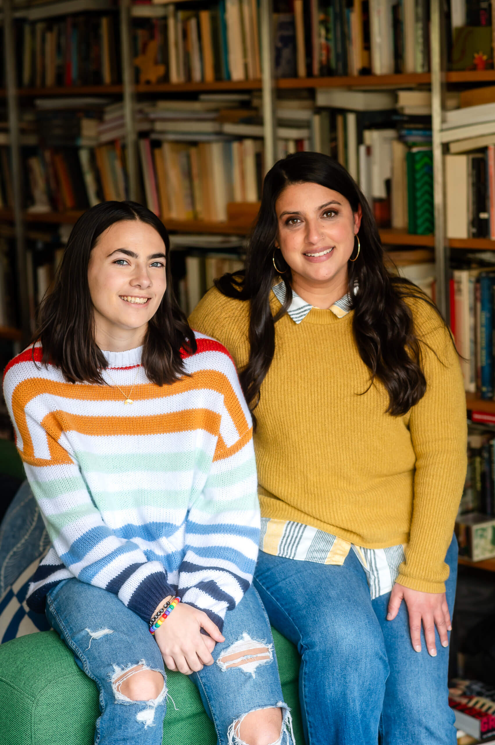 A mentor and LGBTQ+ young person smile in front of a bookshelf.